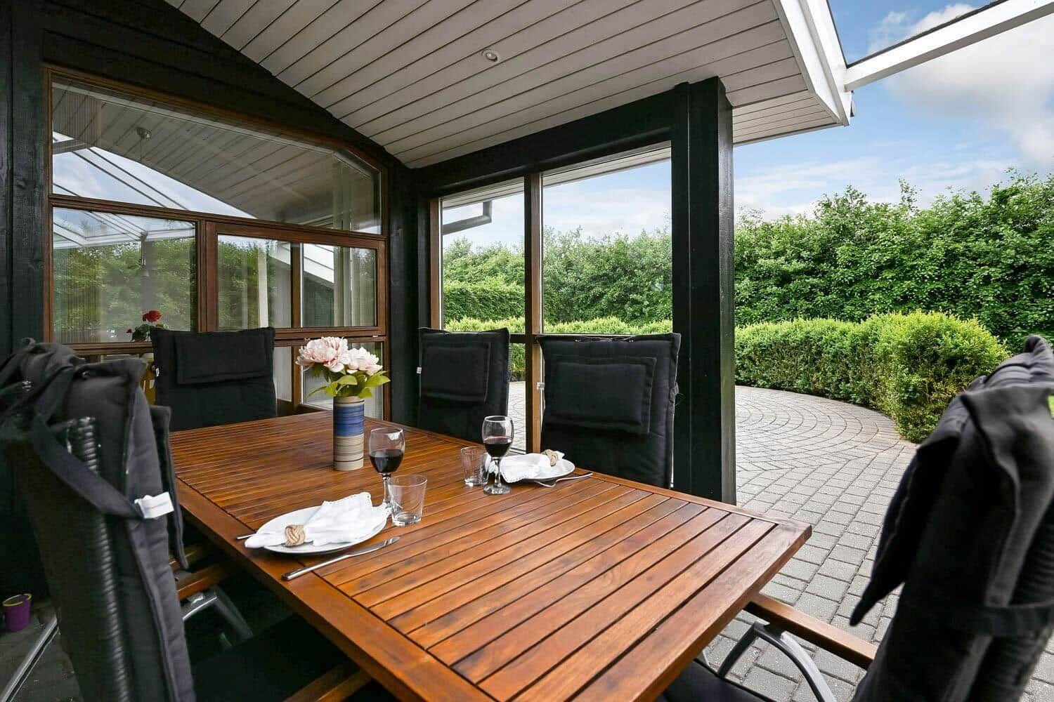 Terrasse mit Holztisch, Stühlen und Blumenarrangement. Blick auf grüne Hecke und gepflasterten Weg.