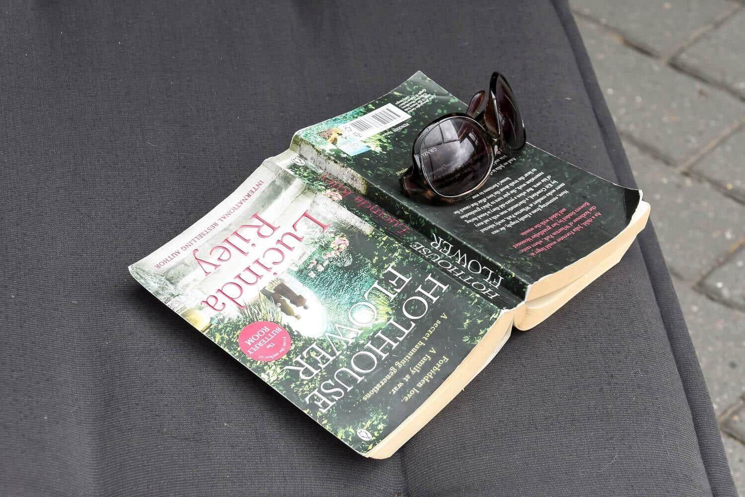 Ein Buch mit Sonnenbrille darauf liegt auf einer dunklen Außentischplatte.