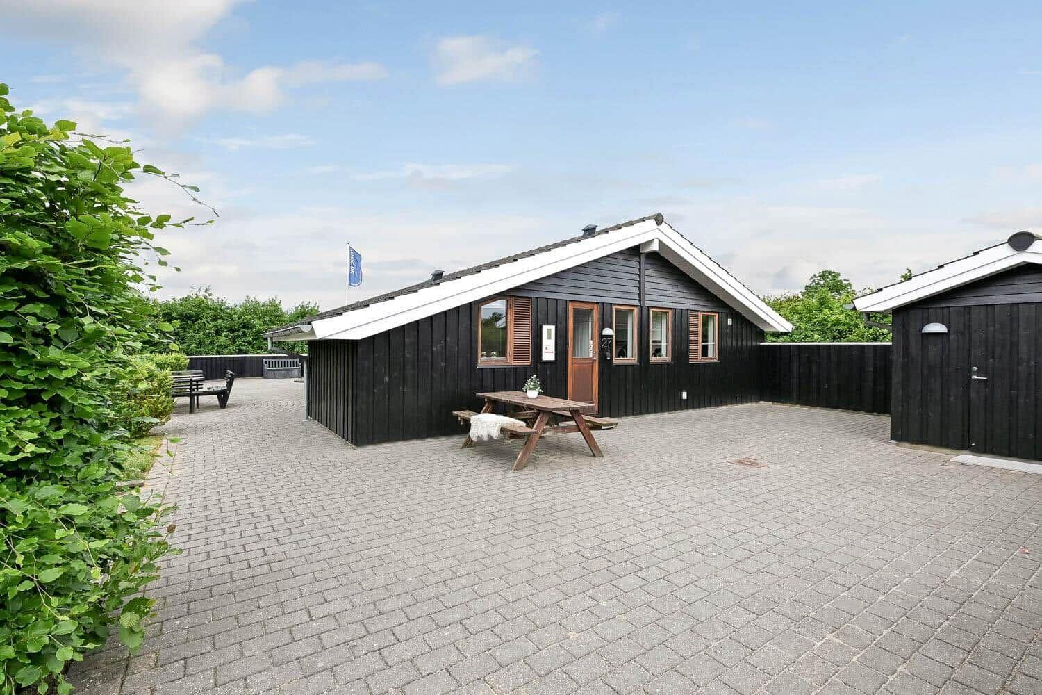Schwarzes Holzhaus mit Terrasse und Picknicktisch. Umgeben von Grün und gepflastertem Hof.