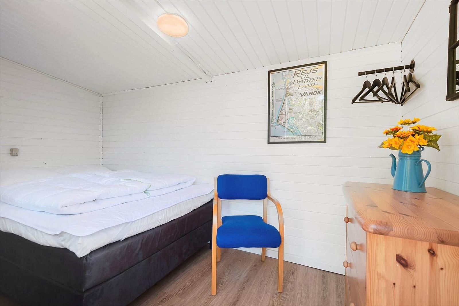 Schlafzimmer mit Bett, blauer Stuhl und Holzkommode. Wandbild und Blumen auf Kommode.