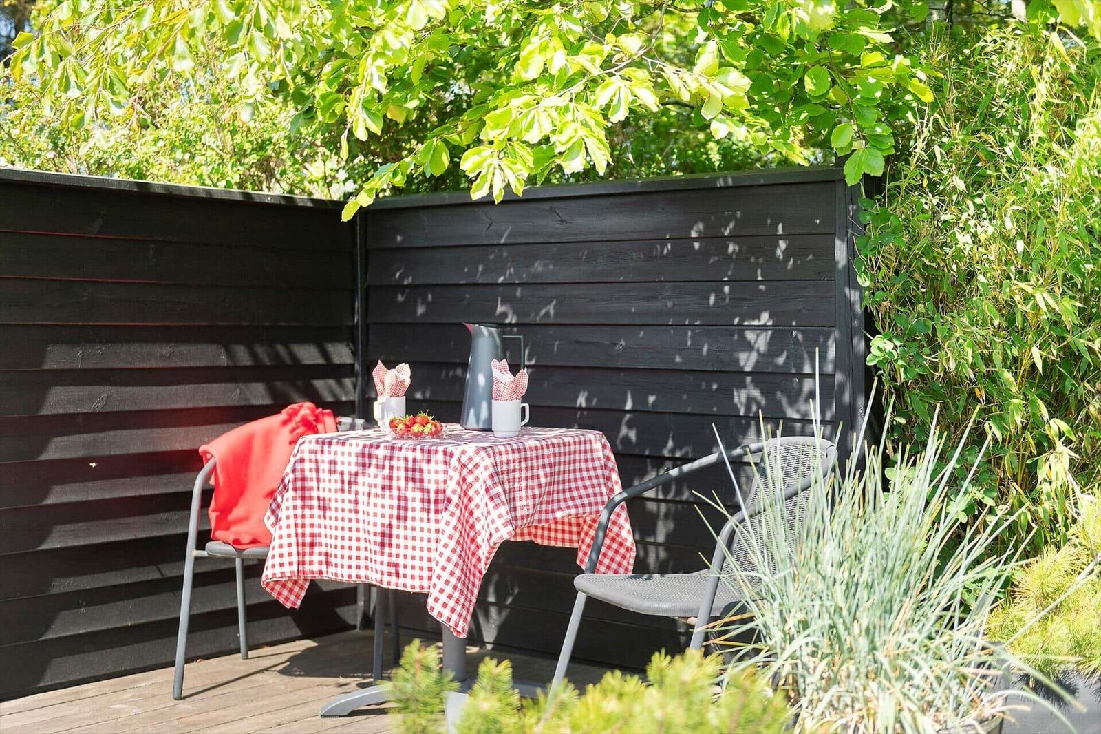 Terrasse mit Tisch, Stühlen und rotem Tischtuch unter Bäumen.