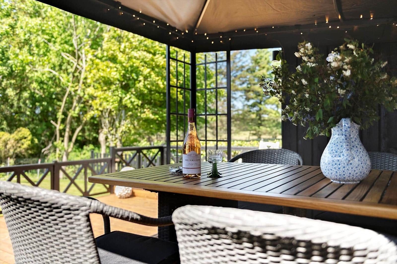 Terrasse mit Tisch, Stühlen, Blumen und Weinflasche unter Markise.