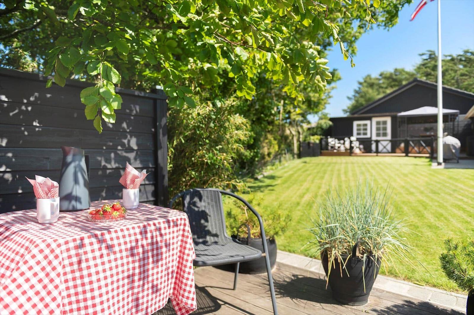 Tisch mit Tischdecke, Stühle und Pflanzen auf Terrasse. Grasfläche und Haus im Hintergrund.