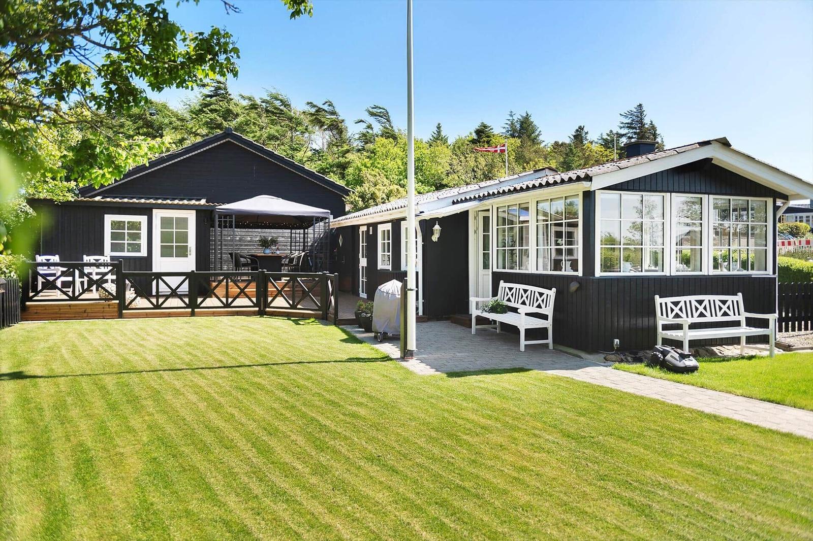 Schwarze Holzhütte mit weißem Geländer, Terrasse und Rasenfläche. Zwei weiße Bänke vor dem Haus.