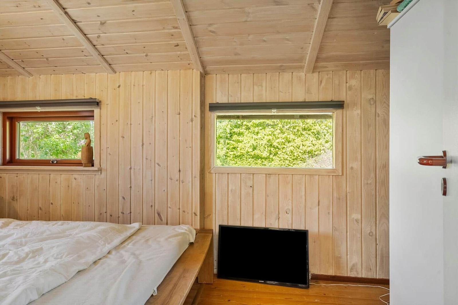 Bedroom with wooden walls, bed, TV, and two windows with view of greenery.