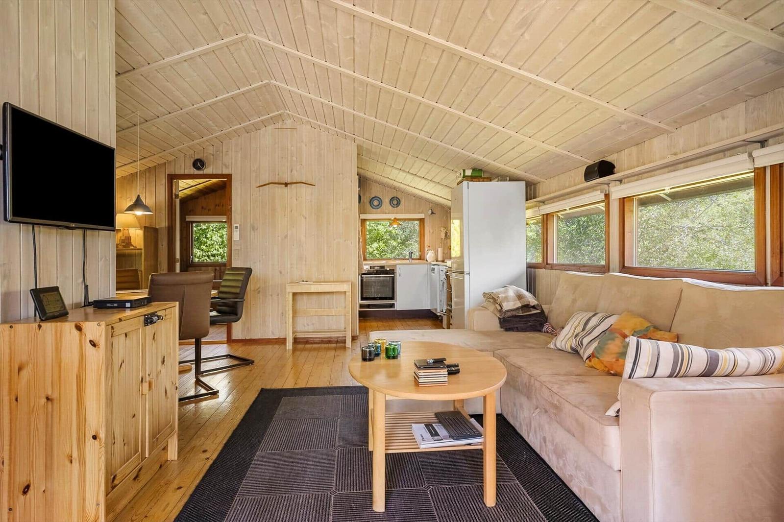 Living area with sofa, coffee table, and TV. Kitchen in background. Wood paneling and large windows.