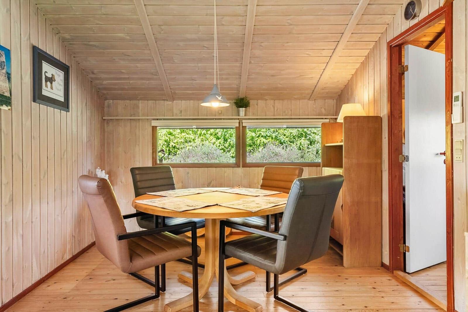 Dining room with round table and four chairs, wooden walls, windows with view of greenery.