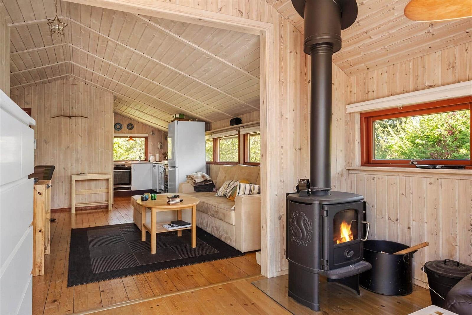 Living area with wood stove, sofa, and kitchen. Wooden walls and windows with view of greenery.