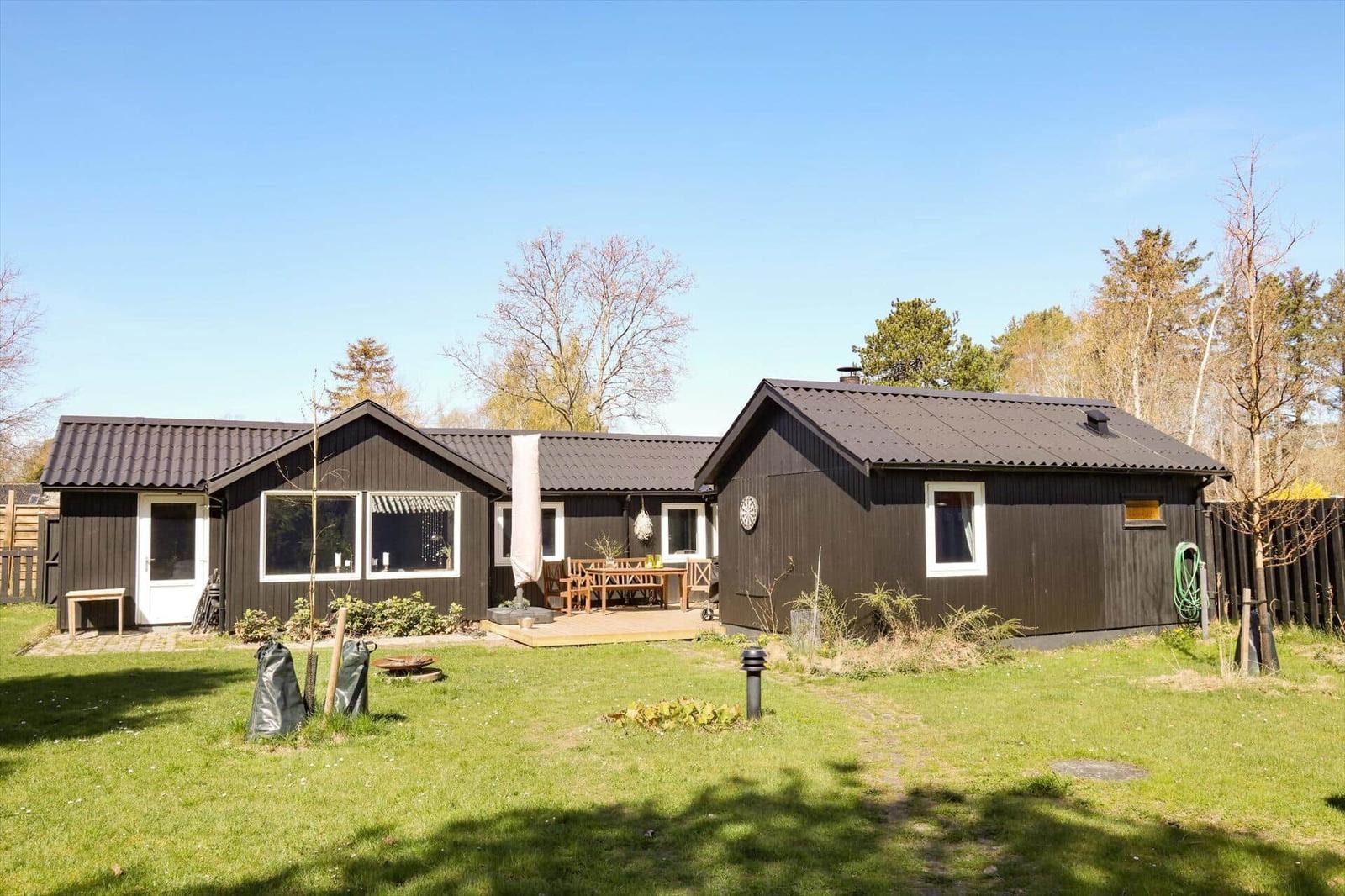 Ein dunkles Holzhaus mit Terrasse und Garten. Umgeben von Bäumen unter blauem Himmel.