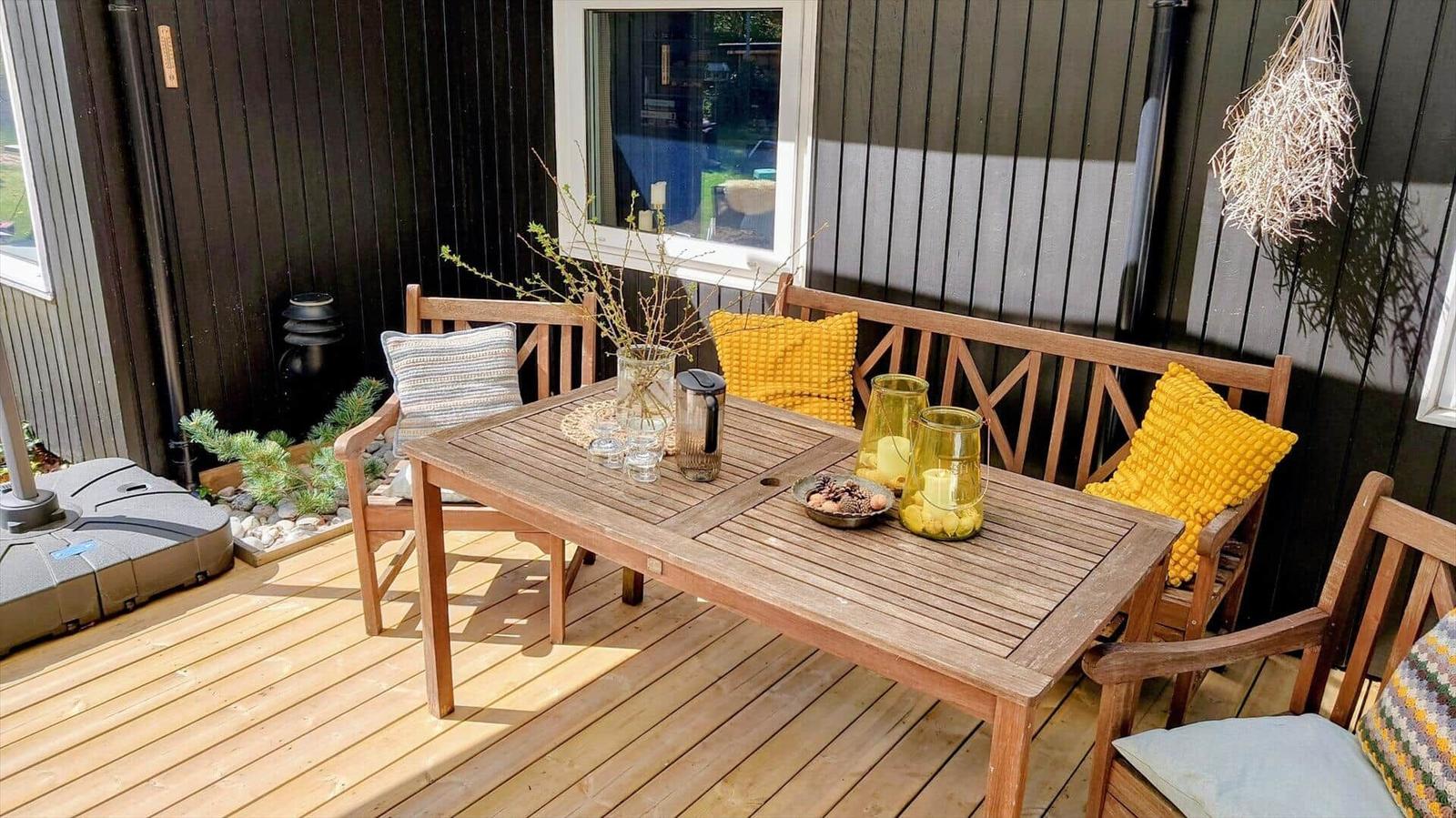 Holzterrasse mit Tisch, Stühlen und Kissen. Fenster und Hängedekoration sind sichtbar.
