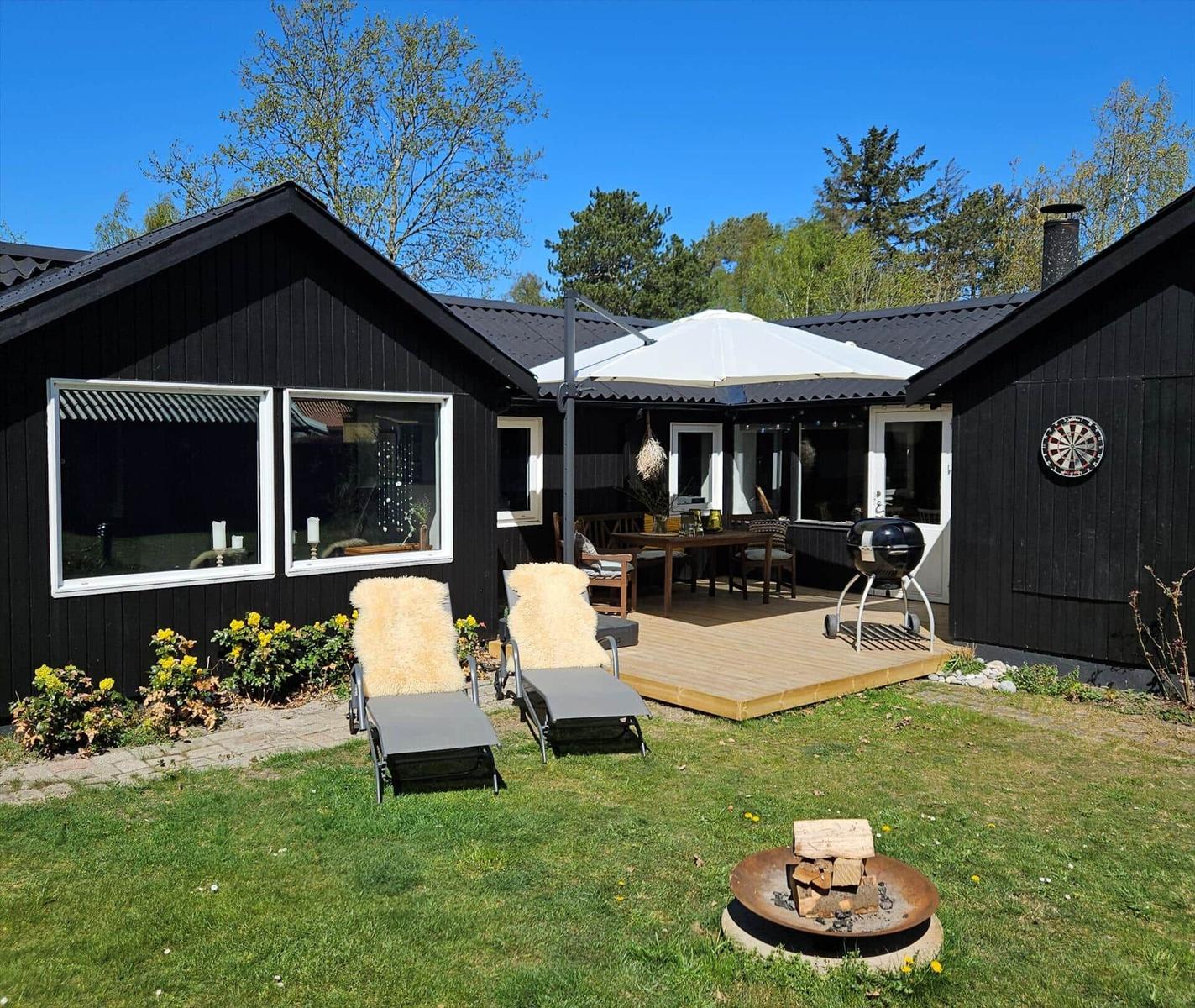 Schwarze Holzhütte mit Terrasse, Liegestühlen und Grill. Feuerkorb und Dartscheibe im Garten.