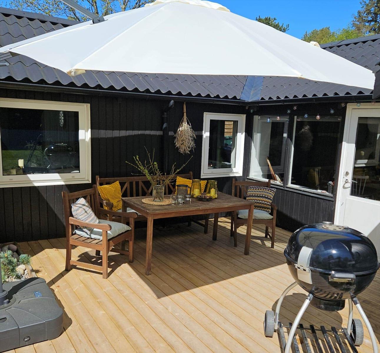 Terrasse mit Tisch, Stühlen, Sonnenschirm und Grill. Holzdecke und schwarze Fassade.