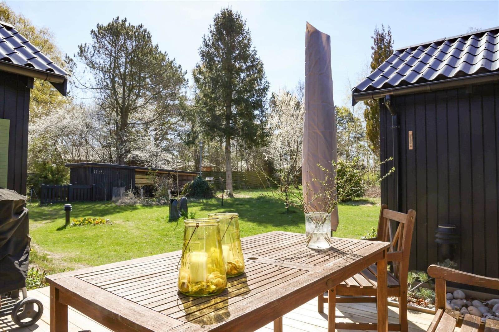 Holztisch mit zwei Stühlen auf Terrasse. Hintergrund: Grasfläche, Bäume und dunkle Holzhäuser.