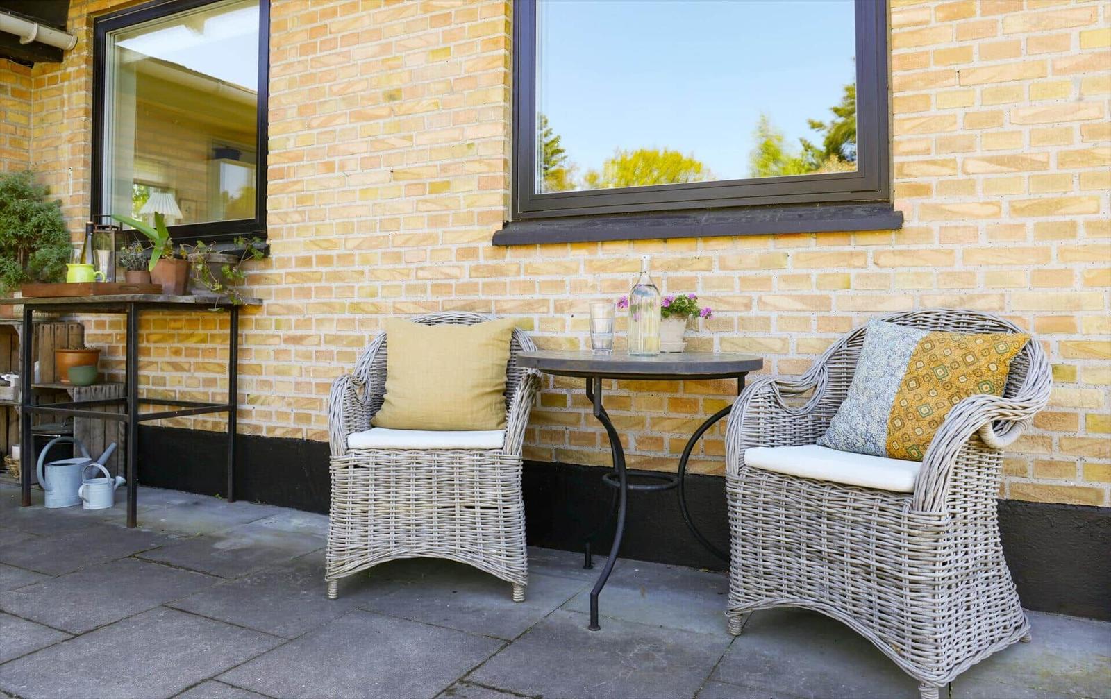 Terrace with two wicker chairs, table, and plants against a yellow brick house.