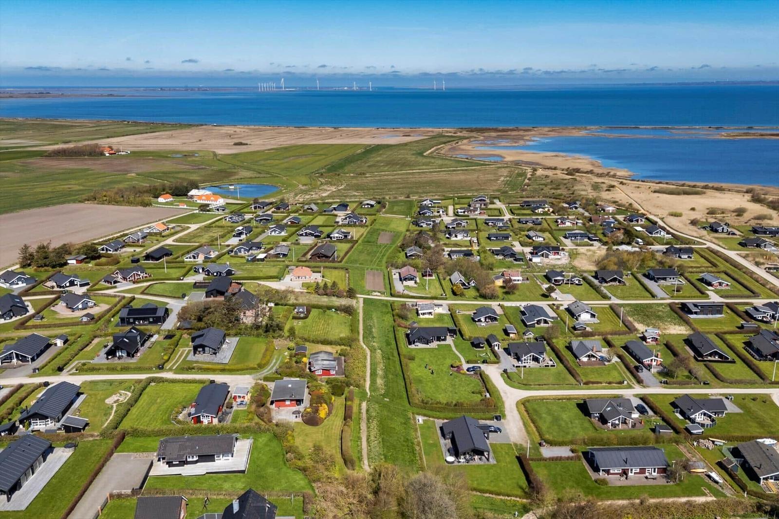 Luftbild eines Ferienortes mit Häusern, Grünflächen und Blick auf Meer und Windkraftanlagen.