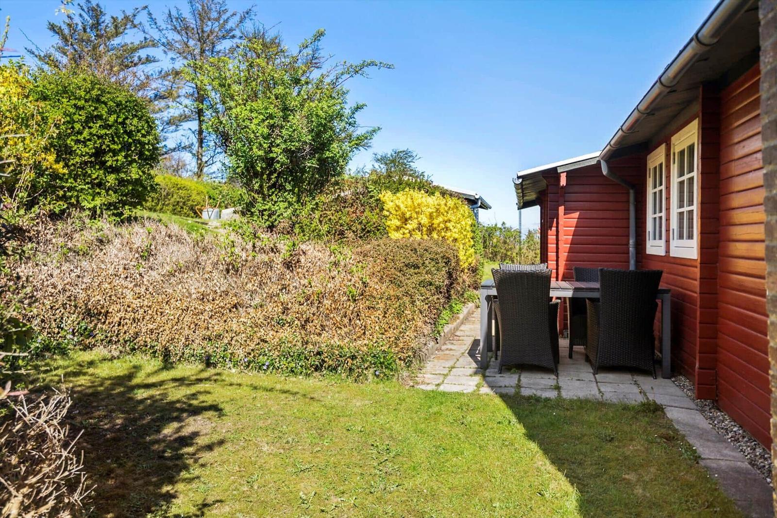 Roter Holzaufbau mit Terrasse, Tisch und Stühlen. Grasfläche und Hecken im Hintergrund.