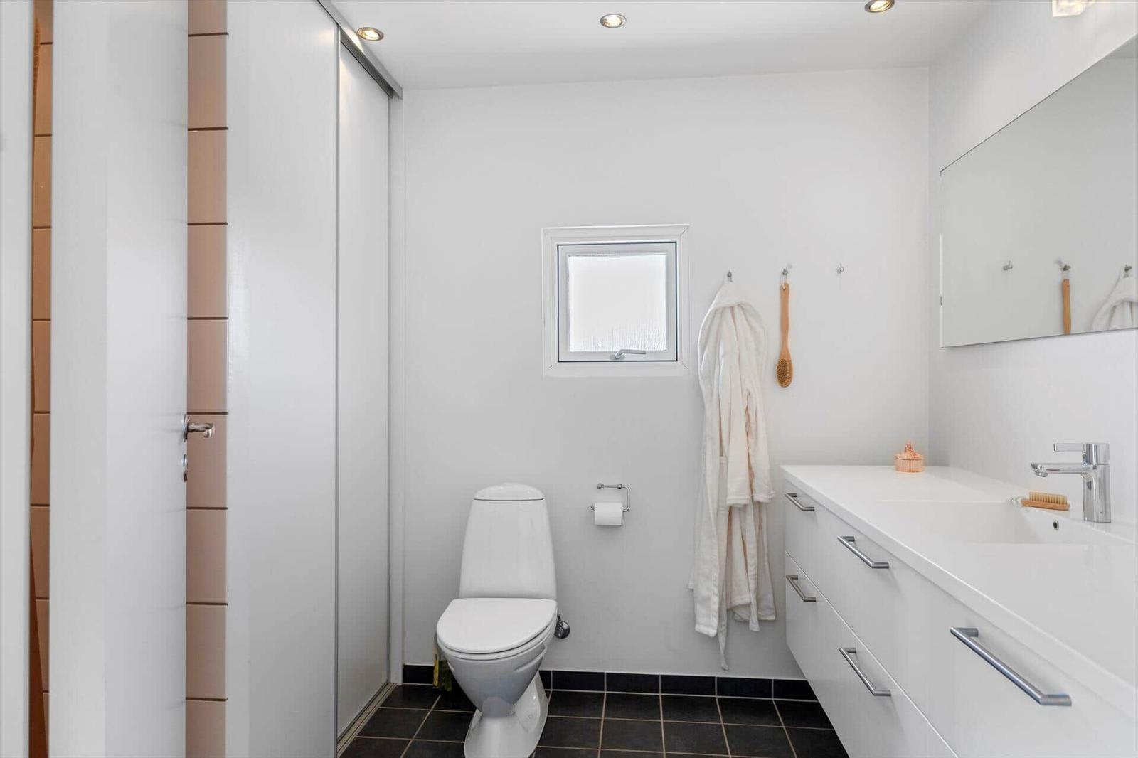 Badezimmer mit Toilette, Waschbecken und Fenster. Weiße Wände und dunkler Boden.