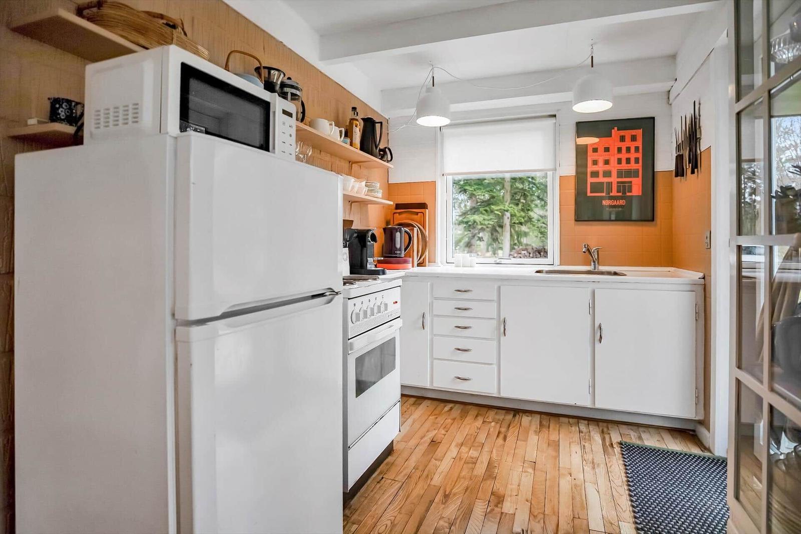 Kitchen with white countertop, refrigerator, stove, and window with view of greenery.