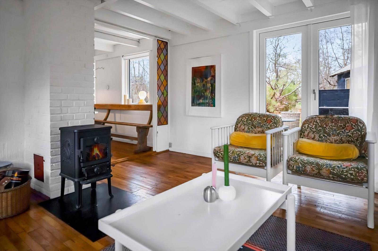 Living room with fireplace, two chairs, and hardwood floor. Windows overlook garden.