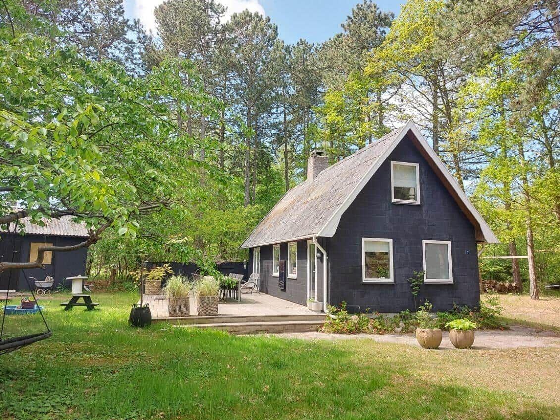 A dark blue wooden house with a roof terrace in the woods. Green lawn and trees surround the house.