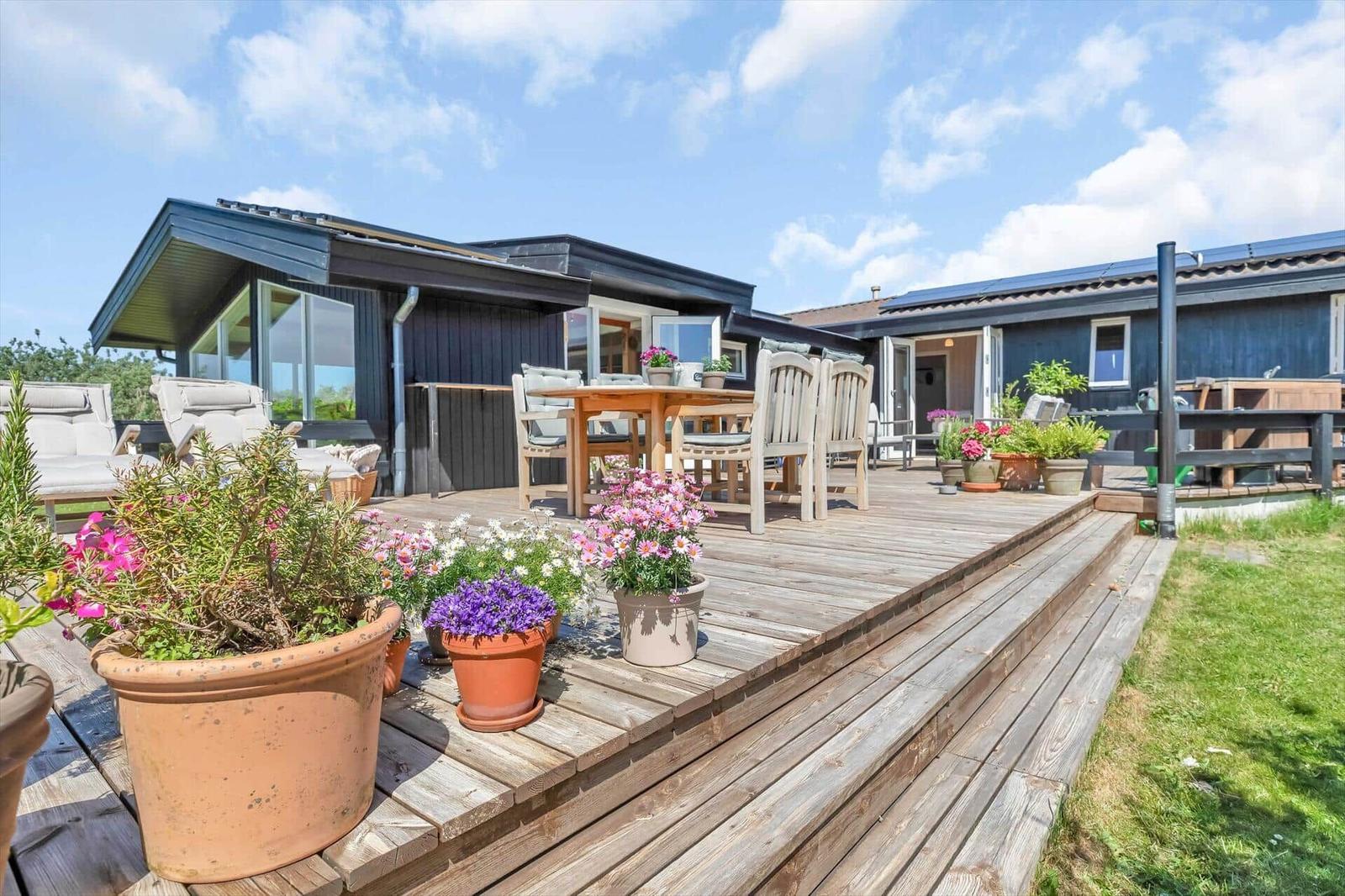 Holzterrasse mit Tisch, Stühlen und Blumenpflanzen vor einem Haus.