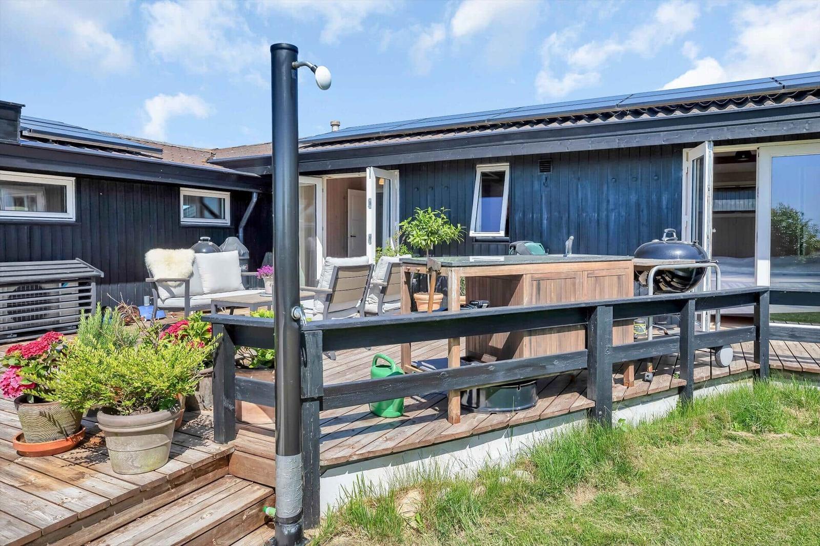 Terrasse mit Grill, Sitzgelegenheiten und Pflanzen vor einem Haus mit dunklem Holzverkleidung.