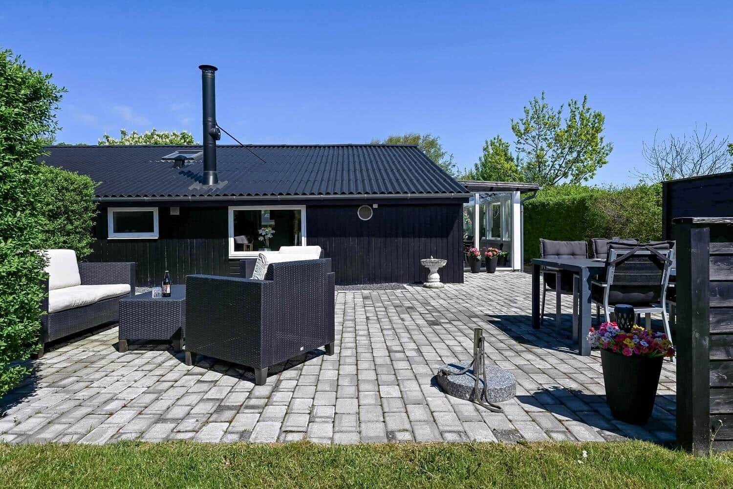 Schwarzes Haus mit großem gepflastertem Hof und Gartenmöbeln unter blauem Himmel.