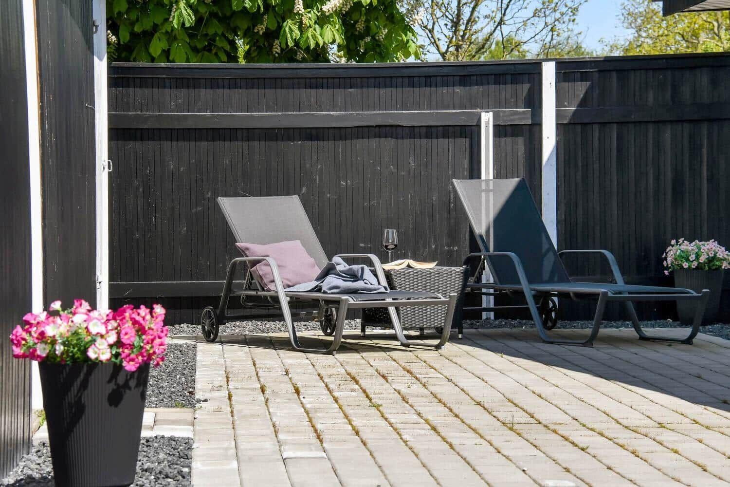 Terrasse mit zwei Liegestühlen, Tisch und Blumenpflanzen vor dunklem Holzzaun.