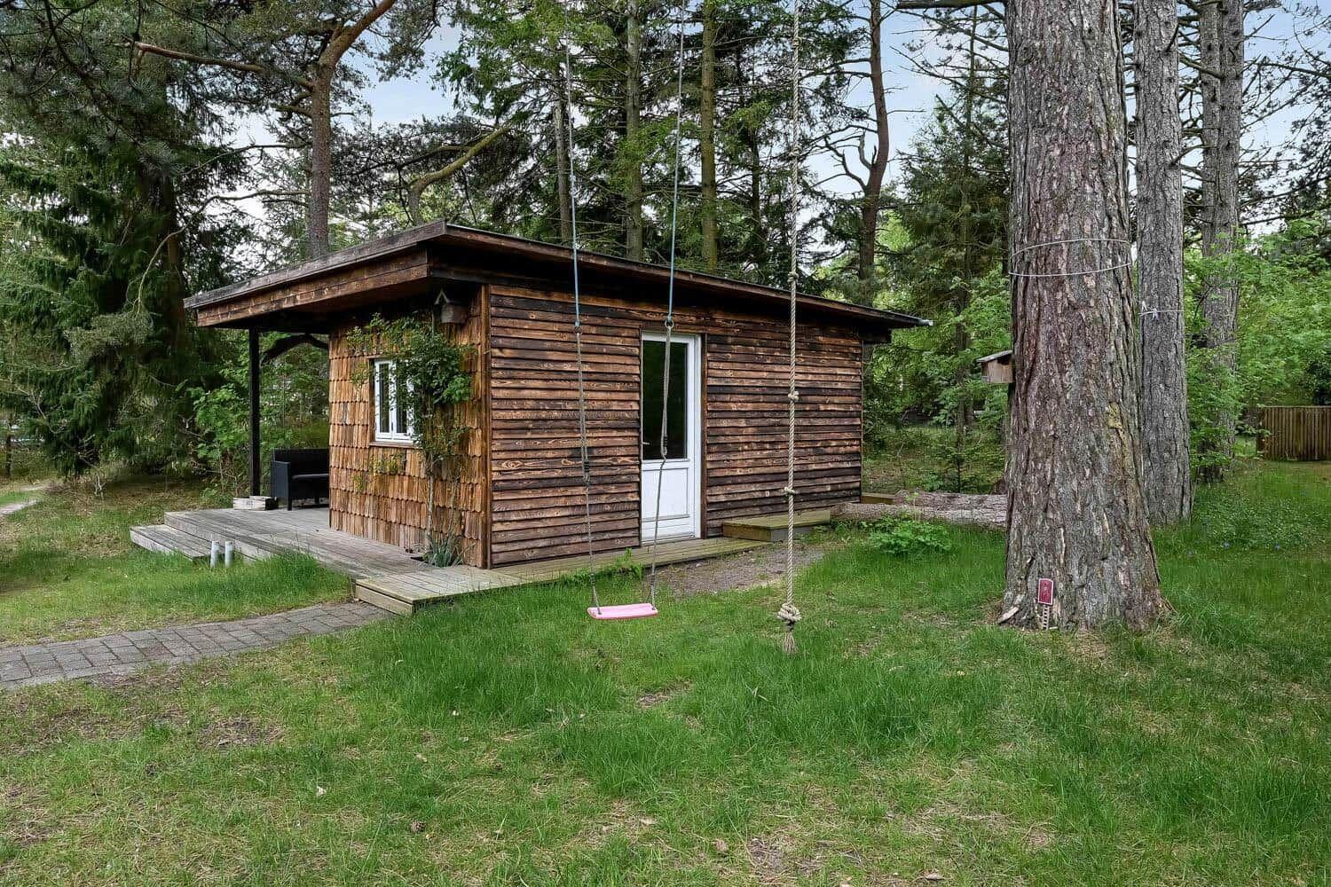 Holzhütte mit Terrasse und Schaukel im Wald. Grünfläche und Bäume umgeben das Gebäude.