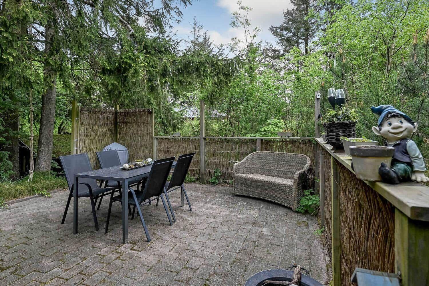 Terrasse mit Sitzgruppe, Tisch und Stühlen, umgeben von Bäumen und einem Zaun.