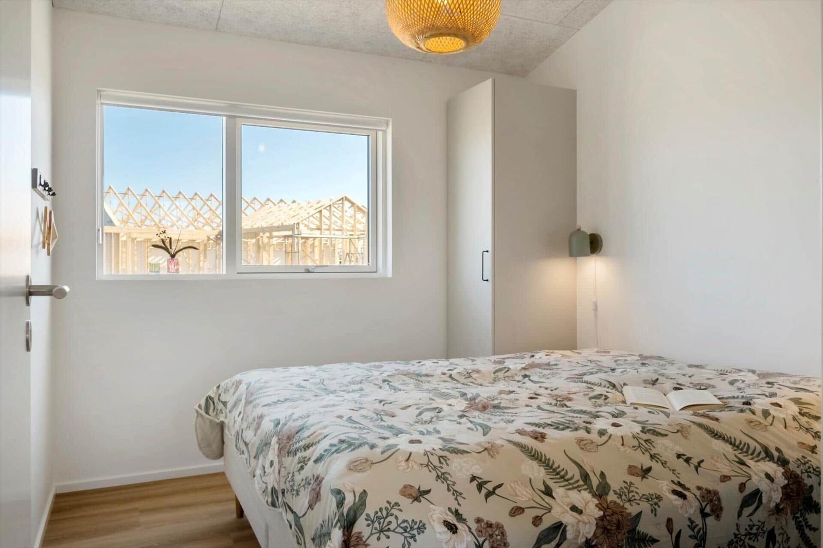 Schlafzimmer mit Bett, Fenster und Deckenleuchte. Draußen sind Holzkonstruktionen sichtbar.