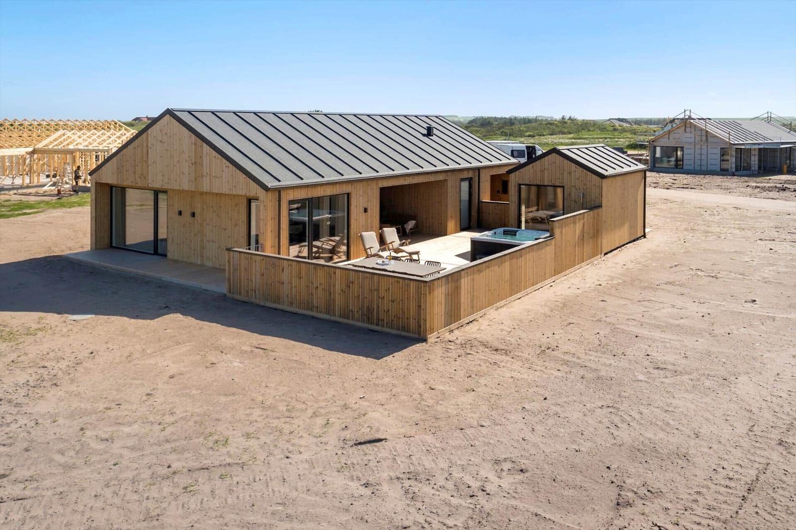 Moderne Holzhütte mit Terrasse und Whirlpool. Umgeben von Baustellen und Sand.