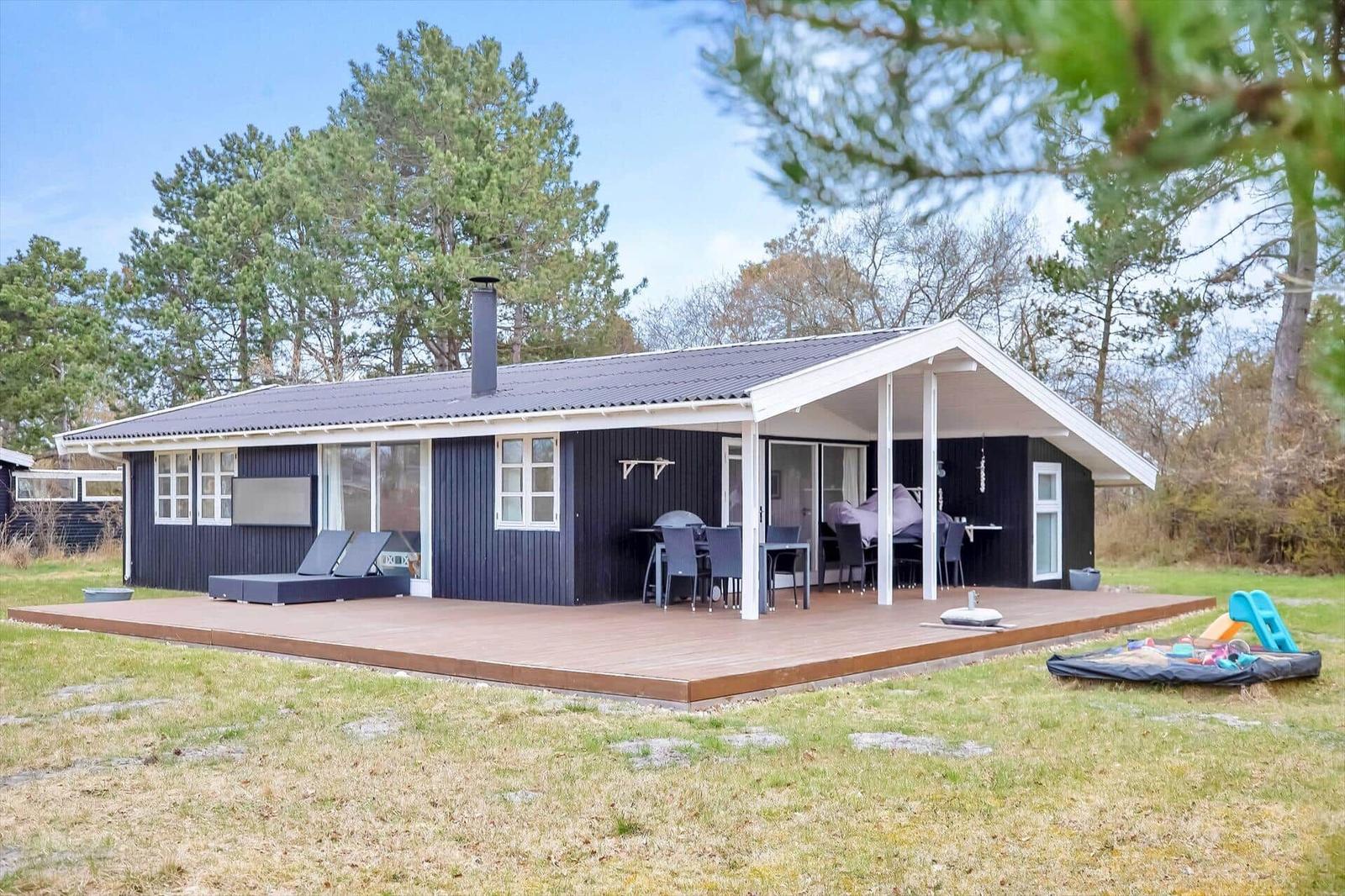 Ein schwarzes Haus mit Terrasse und Garten. Außentisch, Liegen und Spielplatz.