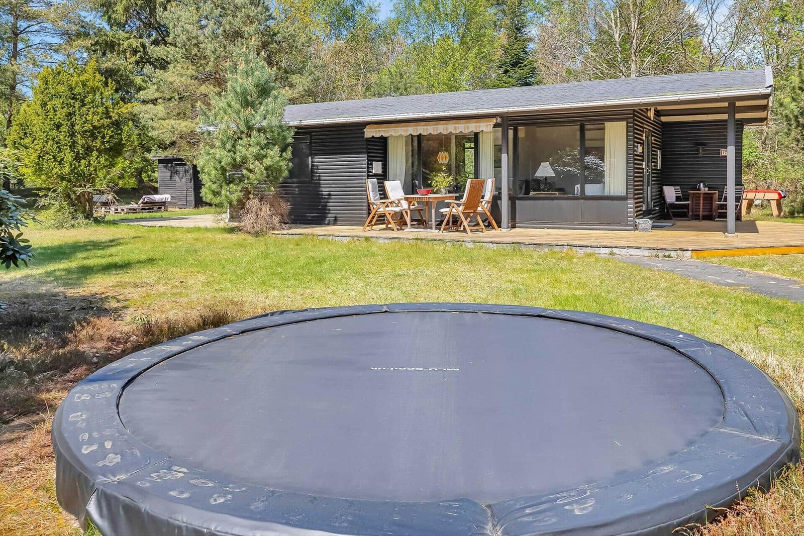 Haus mit Terrasse, Garten und Sprungtisch. Holzmöbel und große Fenster.