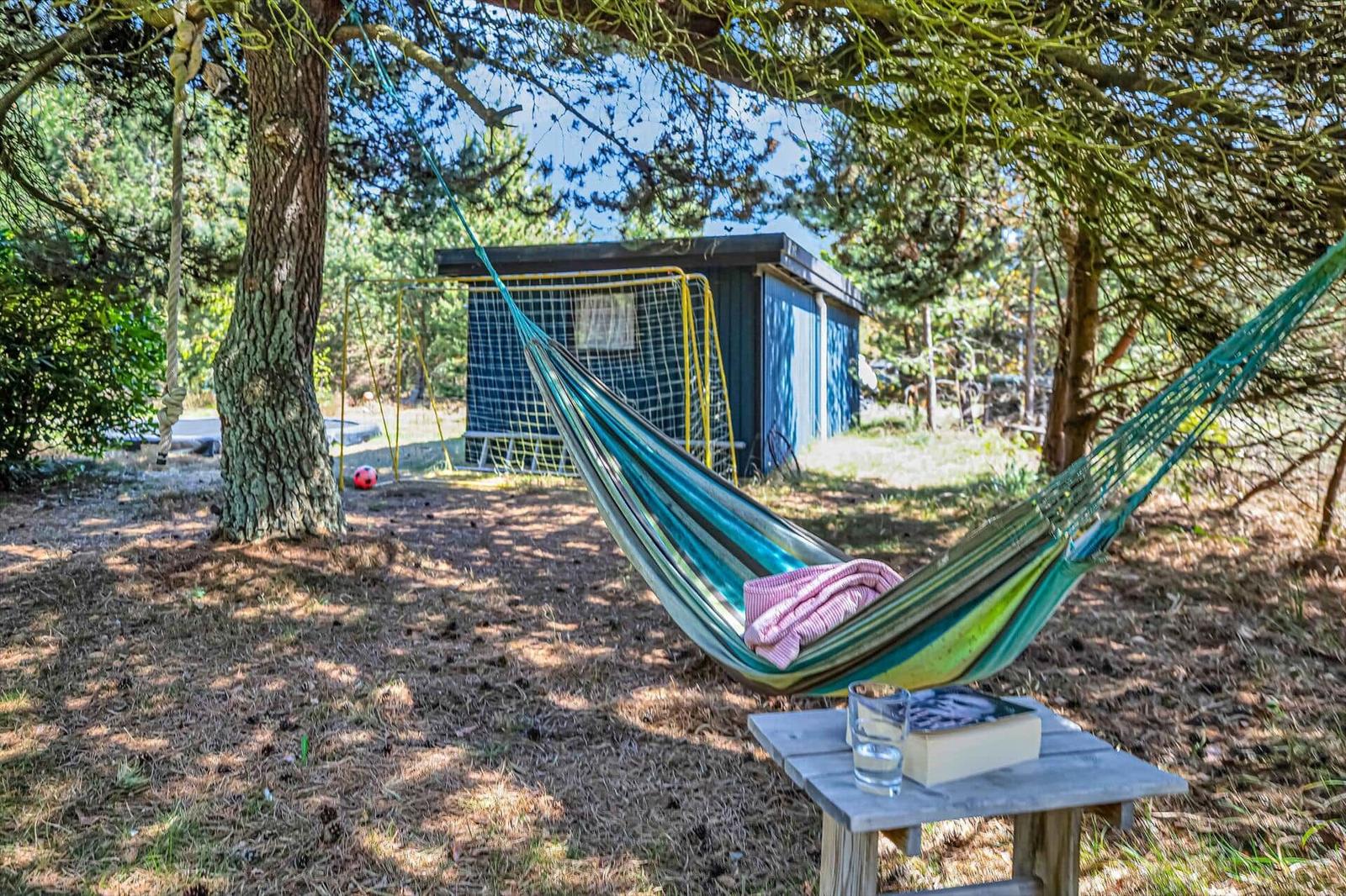 Hammock unter Bäumen mit Tisch, Buch und Glas Wasser. Im Hintergrund ein blauer Schuppen.