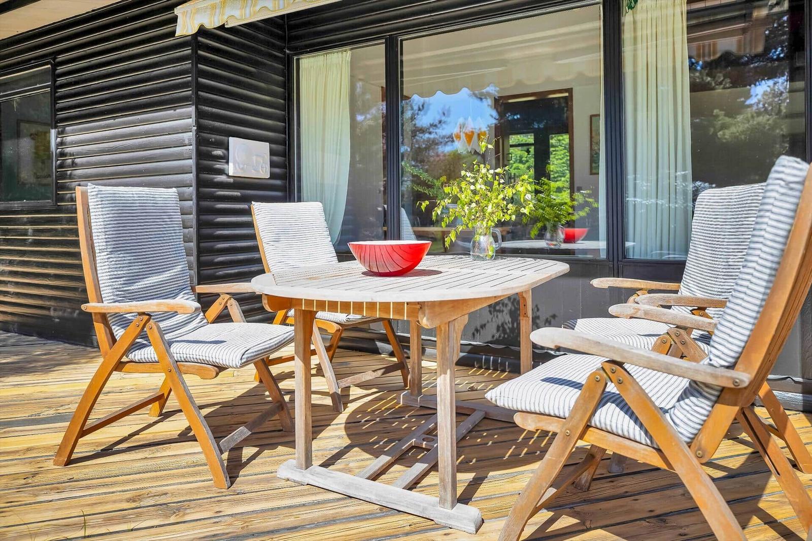Terrasse mit Holztisch und drei Stühlen, roter Schale und Pflanze. Schwarze Holzwand und Glasfront.