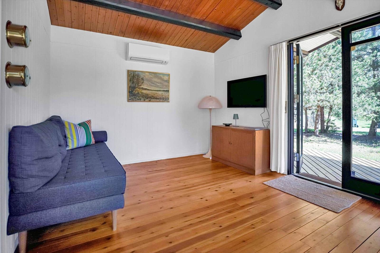 Ein Wohnzimmer mit Sofa, Fernseher und Holzboden. Durchgehendes Glas führt auf eine Terrasse.