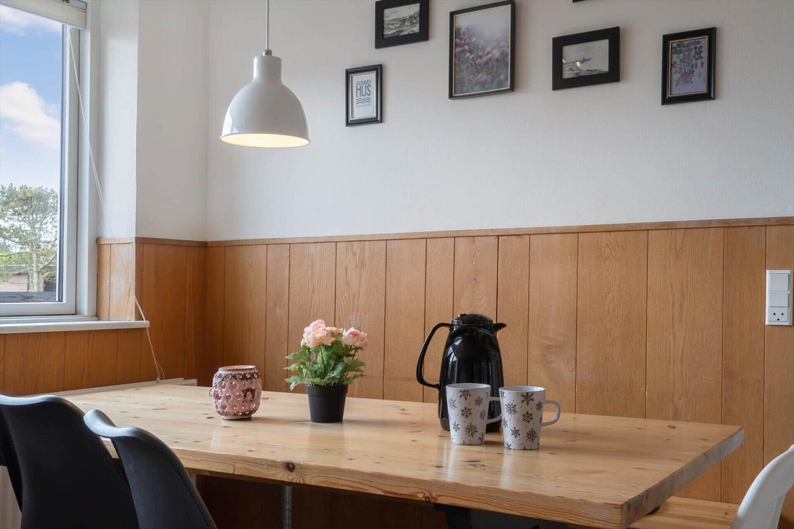 Wooden table with coffee mugs, pot, and flowers against wooden wall and window.