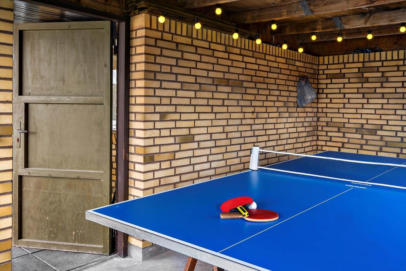 Ping pong table with paddles and ball in a room with brick wall and string lights.