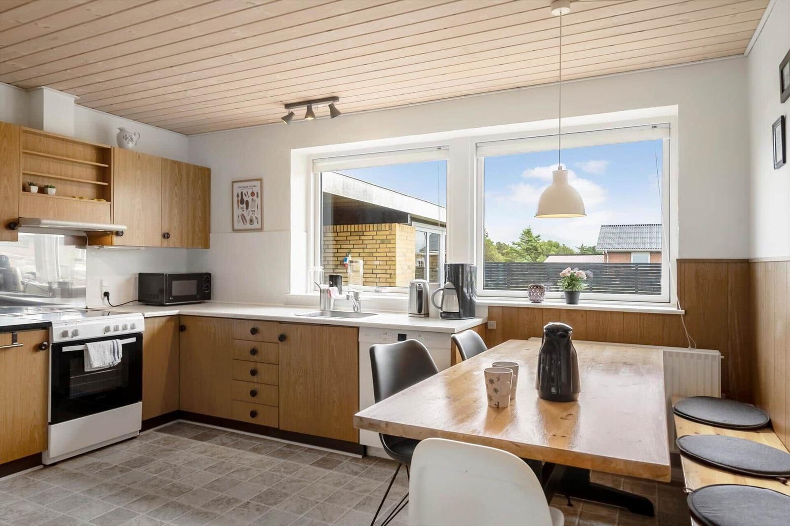 Kitchen with wooden cabinets and dining table. Windows overlook the outdoor area.