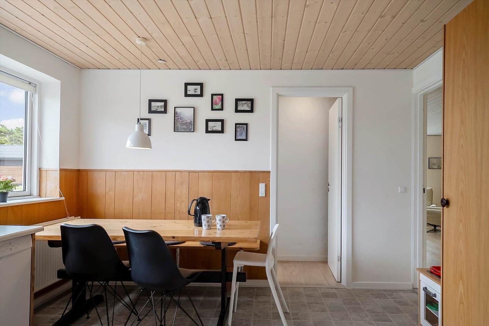 Dining area with wooden table, black chairs, and wall pictures. Wooden ceiling and window view.