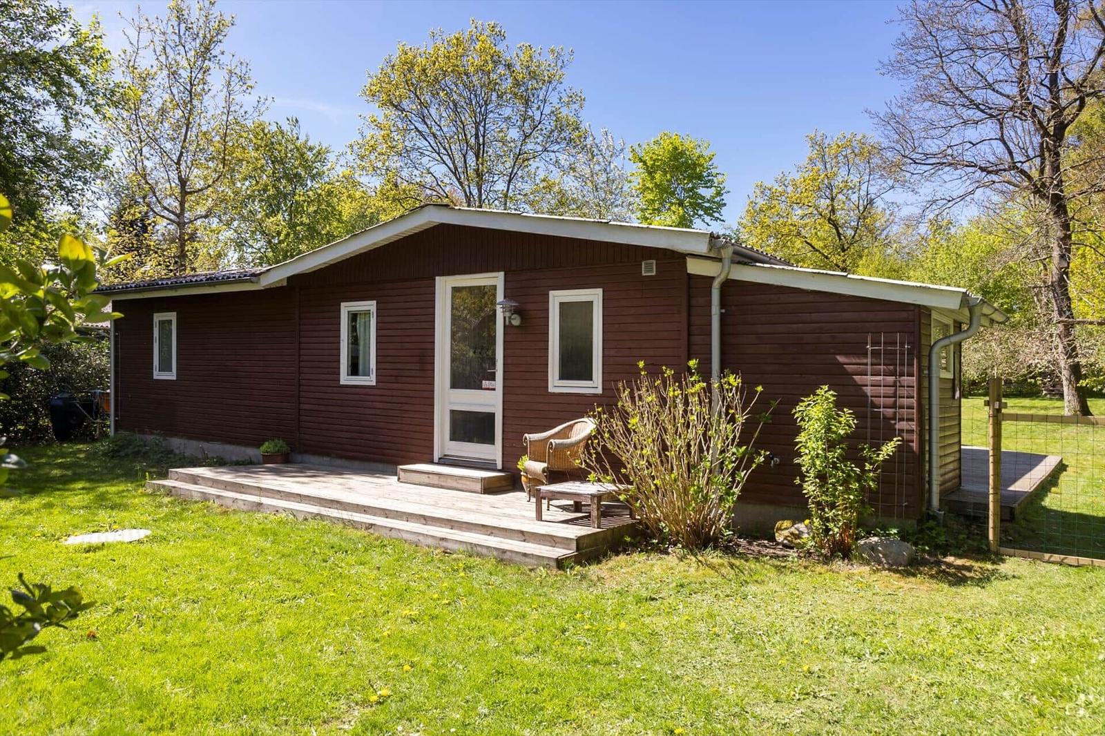 A brown wooden house with a terrace, garden, and trees in the background.