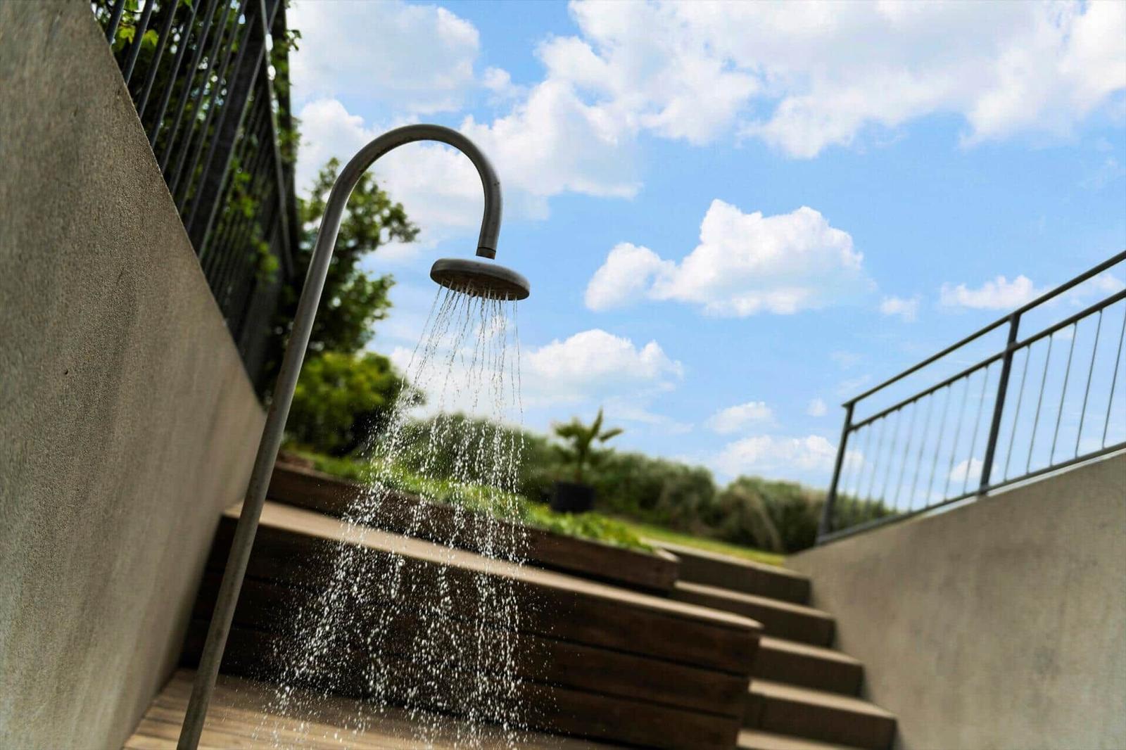 Außendusche mit Wasserstrahl auf Holzstufen unter blauem Himmel mit Wolken.