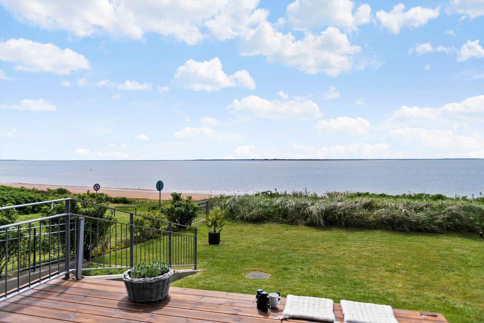 Terrasse mit Blick auf Meer und Strand. Grasfläche und Metallgeländer sichtbar.