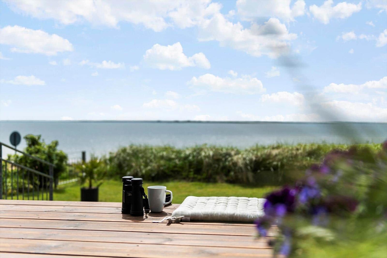 Holztisch mit Kissen, Tasse und Fernglas auf Terrasse mit Blick auf Wasser und Himmel.