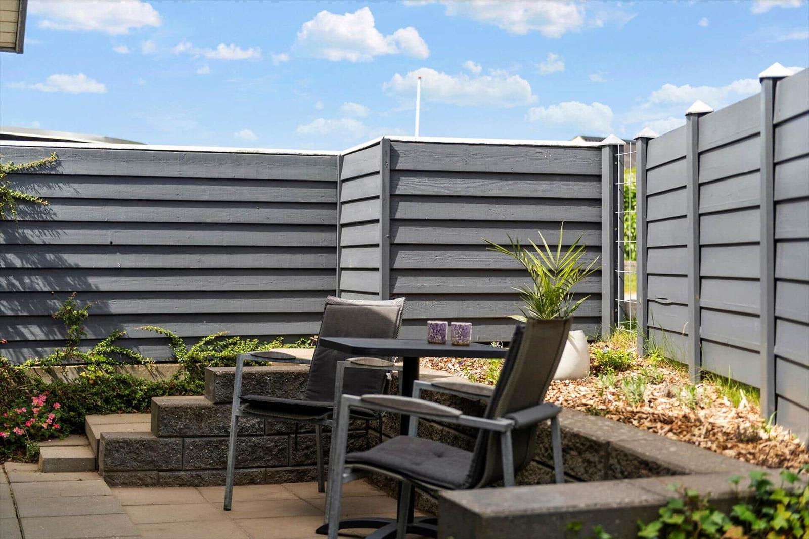 Terrasse mit Tisch, zwei Stühlen und Pflanzen. Umgeben von grauem Holzzaun.
