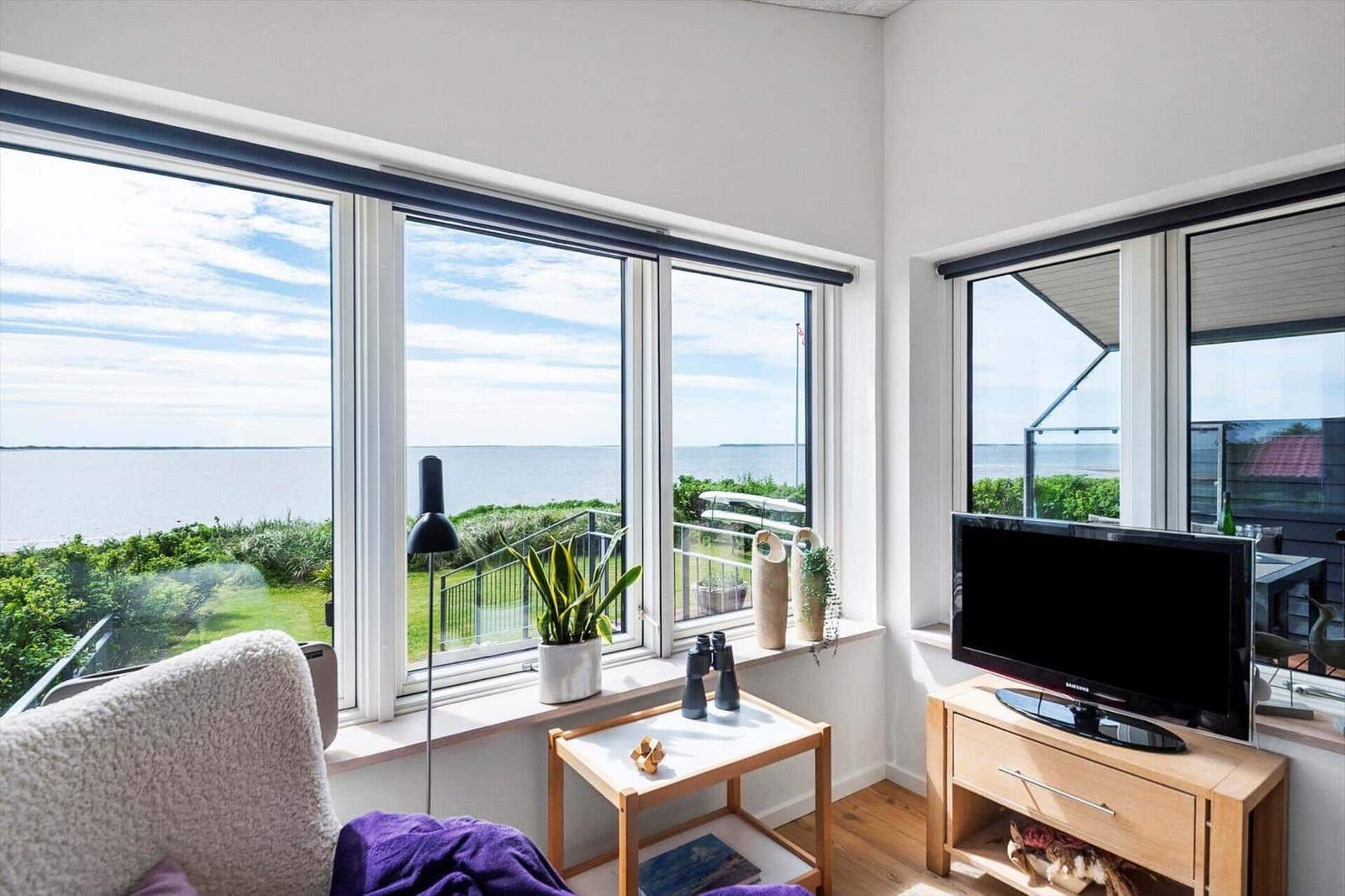 Wohnzimmer mit Meerblick durch große Fenster, Fernseher und Pflanzen auf Fensterbank.