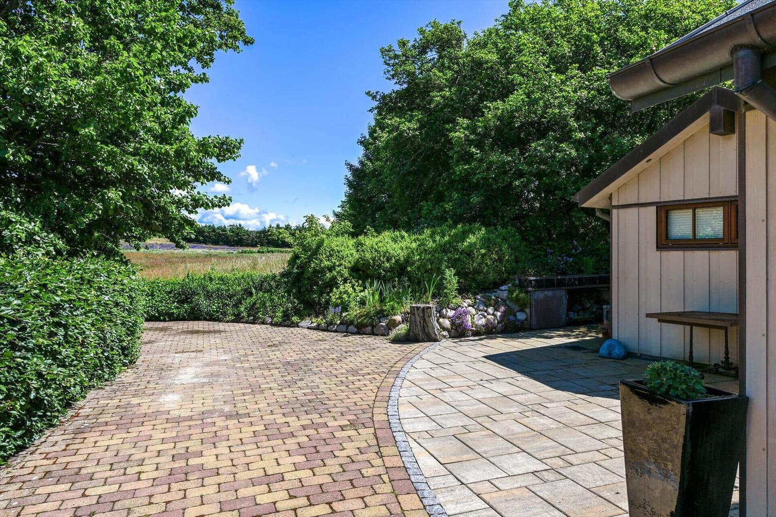 Paved path leads to a wooden cabin with garden and forest view.