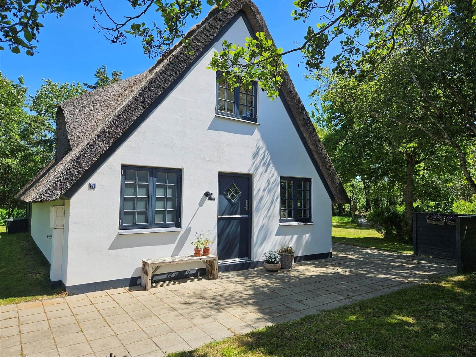 Wohnhaus mit Strohdach, weißen Wänden und dunklen Fenstern. Vor dem Haus befindet sich eine Terrasse.