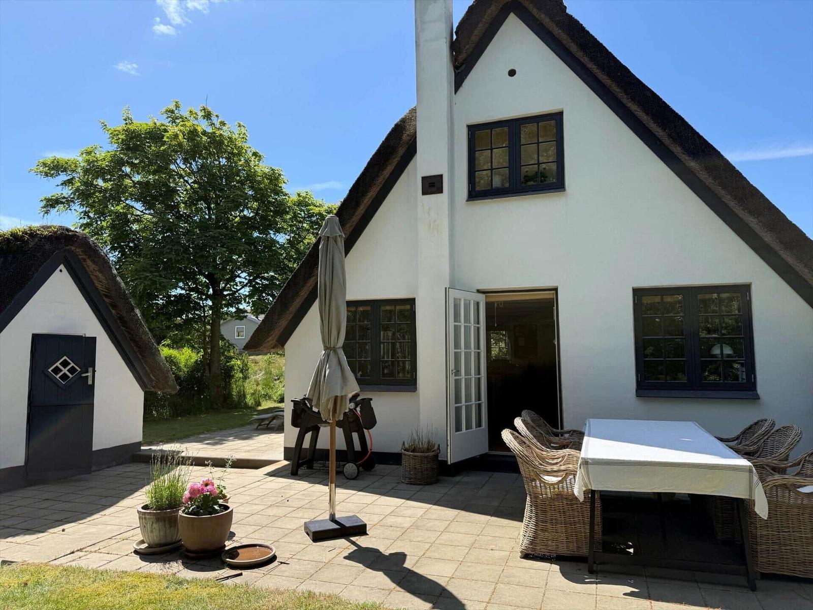 Wohnhaus mit strohgedecktem Dach, Terrasse mit Tisch und Stühlen, Gartentüren und Pflanzen.